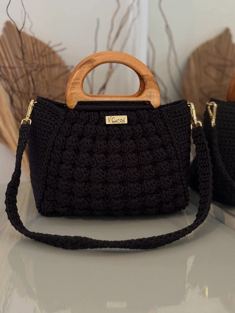A black crocheted handbag with a round wooden top handle and a matching shoulder strap, featuring gold-tone hardware and a small metal brand plaque reading &