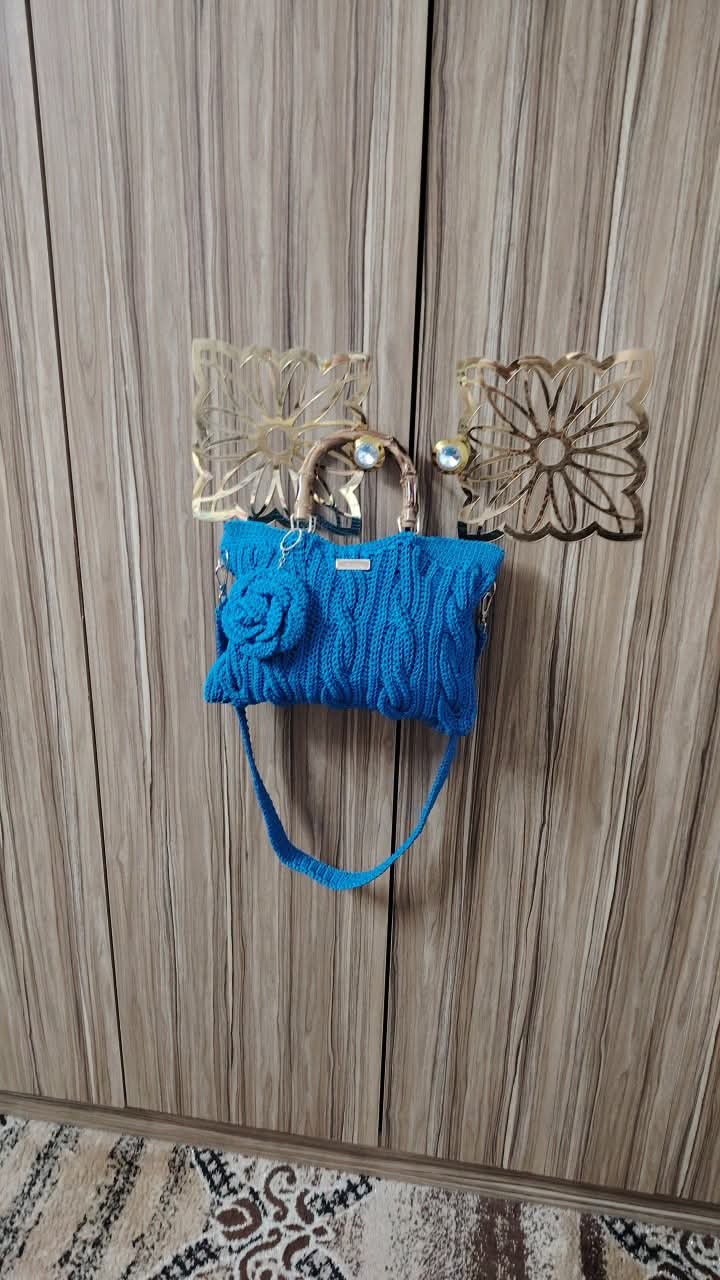A bright blue knitted handbag with a floral detail on the front, hanging from decorative metal hooks on a wood-paneled wall.
