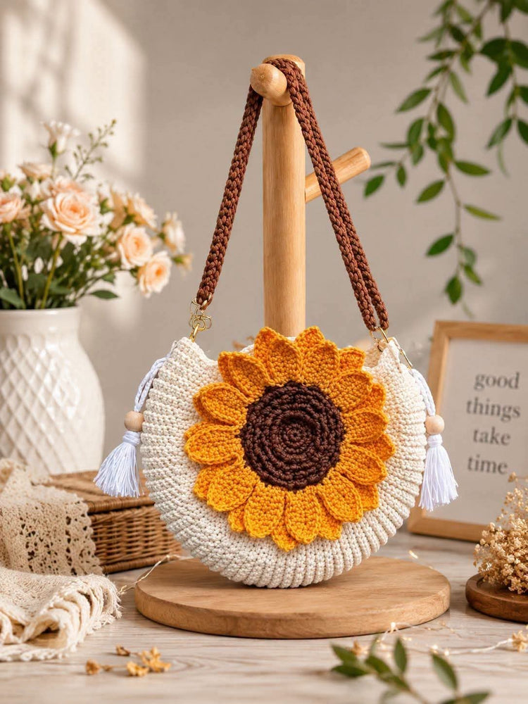 A crocheted sunflower-shaped shoulder bag with a brown braided strap, displayed on a wooden stand. The bag features a cream-colored crochet body with a large yellow and brown sunflower applique on the front and white tassel details on the sides.