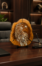 A modern office interior with a large wooden desk, black leather office chair, shelving, plants, and a decorative round wall piece featuring a stylized bird head in warm brown, gold, and cream tones mounted on the wall.

