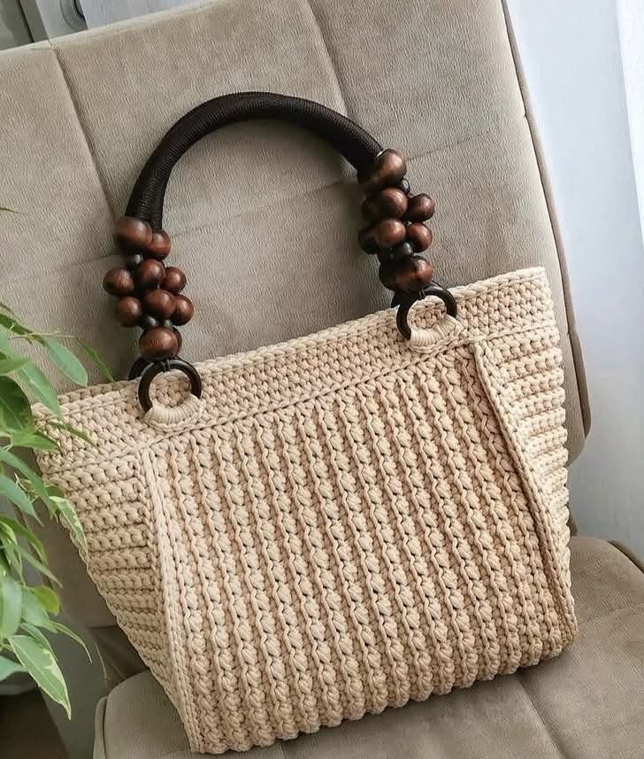 A beige crocheted tote bag with dark brown wooden handles and matching wooden bead accents, resting on a beige upholstered chair.

