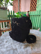 A black crocheted handbag with a matching detachable shoulder strap, displayed on a white faux fur surface with a green picket fence and potted plants in the background.

