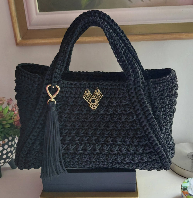 A black crocheted tote bag with two top handles, a gold heart-shaped metal emblem on the front, and a matching black tassel charm, displayed on a stand.

