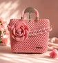 A pink crocheted handbag with a large 3D flower embellishment on the front, a braided handle, and a small metal brand plaque near the bottom right corner, set against a soft pink background with decorative rose petals.

