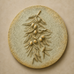 A round textured wall plaque featuring a raised olive branch with small olive fruits, shown against a matching beige background.

