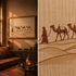 A split view of a framed desert camel caravan artwork hanging in a warm, rustic room on the left, and a close-up of the textile-style artwork on the right. The scene shows tan and brown camels and figures on a beige ground, with the room featuring warm wood tones, patterned textiles, and lanterns.

