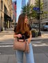A woman carrying a woven beige handbag with a tassel and chain strap, standing on a city street corner.

