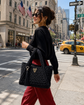 A woman walking on a city street carrying a black woven tote bag with a gold-tone logo and tassel detail. The background shows a busy urban street scene with buildings, flags, a yellow taxi, and a One Way sign.

