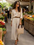 A woman walking through a farmers market wearing a cream-colored button-down shirt and matching wide-leg trousers, accessorized with a brown belt, sunglasses, and a woven straw handbag.

