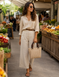 A woman walking through a farmers market wearing a cream-colored button-down shirt and matching wide-leg trousers, accessorized with a brown belt, sunglasses, and a woven straw handbag.

